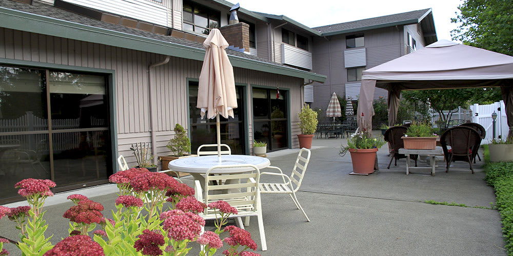 A cozy patio area with a table, chairs, and umbrella surrounded by potted plants in front of an apartment building.