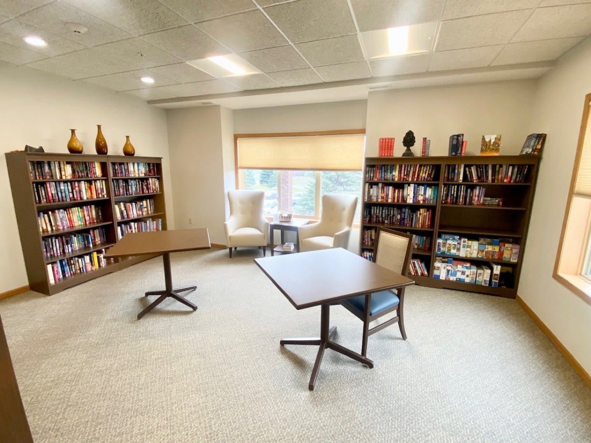 A cozy library space with bookshelves, tables, and chairs arranged near a window.