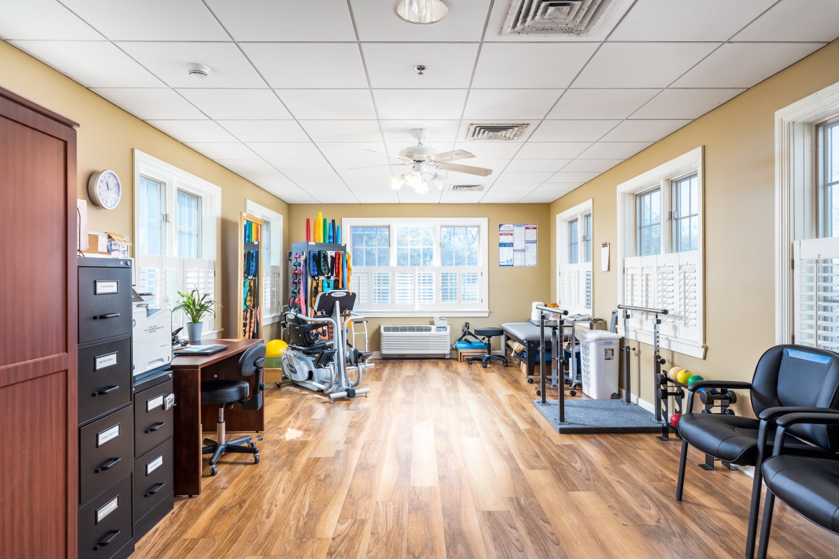 A spacious rehabilitation room filled with therapy and exercise equipment, featuring bright lighting and wooden flooring.