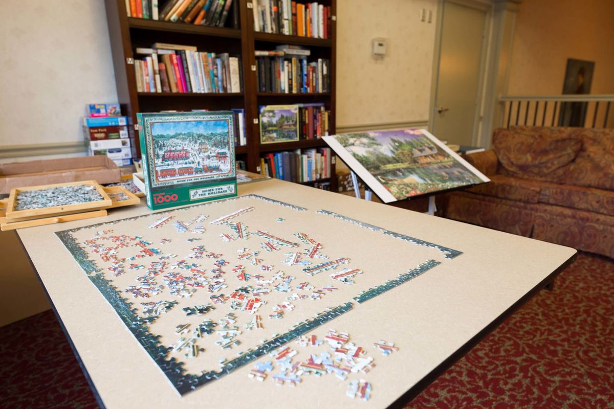 A library setting with a partially completed jigsaw puzzle on a table surrounded by bookshelves.