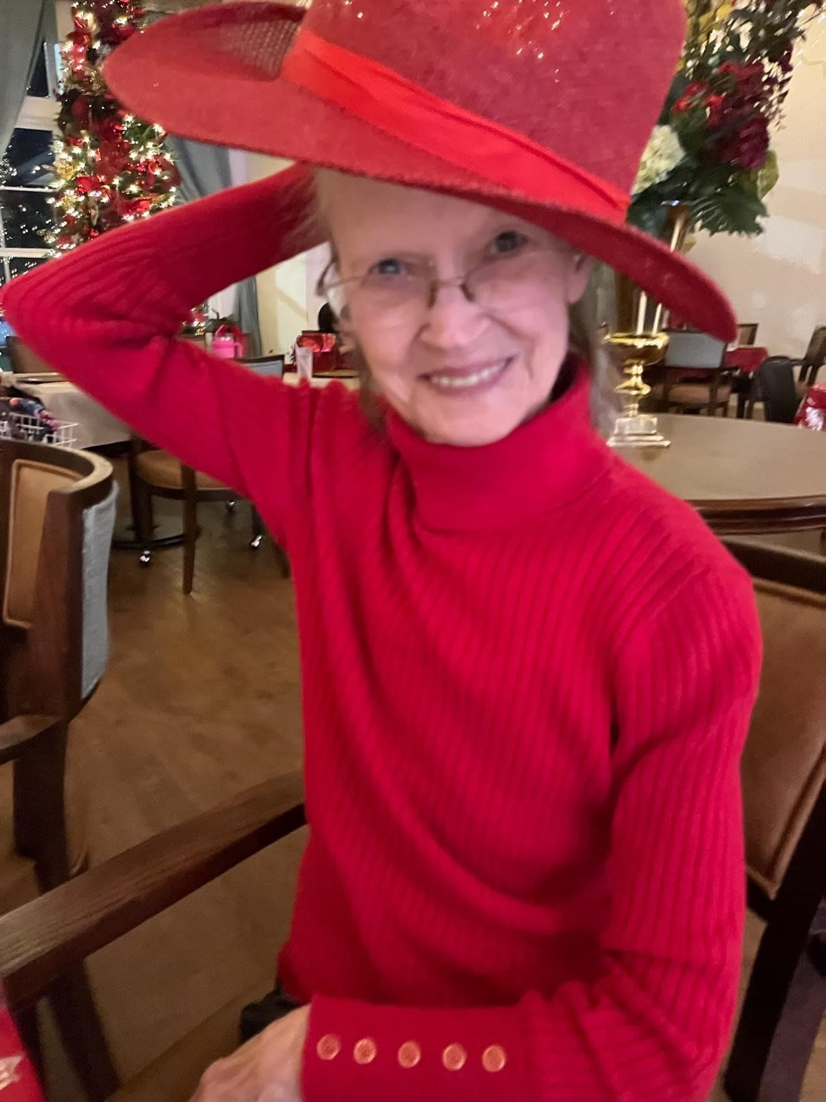 A smiling woman in a red dress and hat, sitting indoors with holiday decorations in the background.