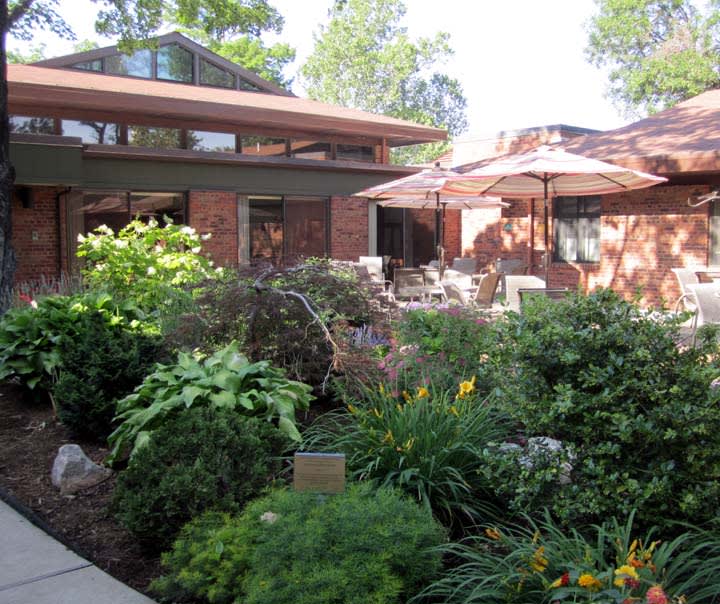 A beautiful garden with lush greenery surrounds a brick house featuring a cozy patio with outdoor furniture and umbrellas.