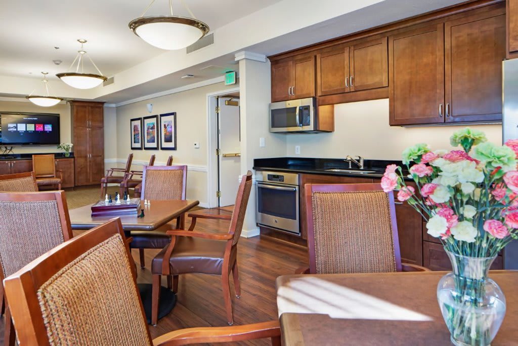 A cozy dining area with wooden furniture and a kitchenette in the background, featuring flowers on the table.