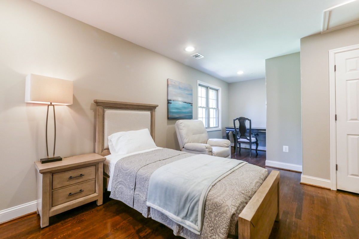 A cozy bedroom with a single bed, nightstand, lamp, armchair, desk, and window.
