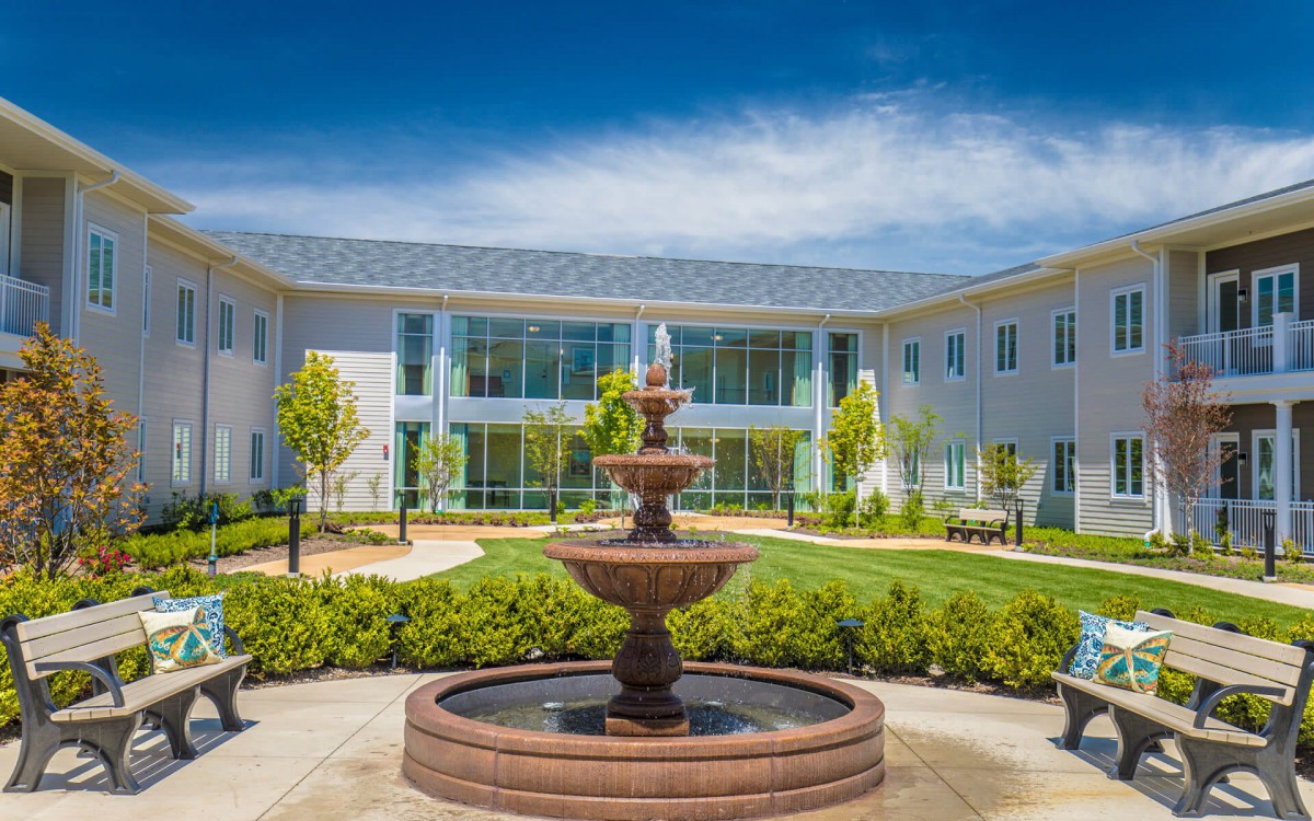 A beautiful courtyard featuring a central fountain surrounded by greenery and modern architecture under a clear blue sky.