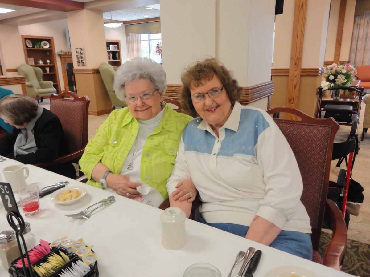 Two elderly women smiling and holding hands while sitting at a dining table indoors.