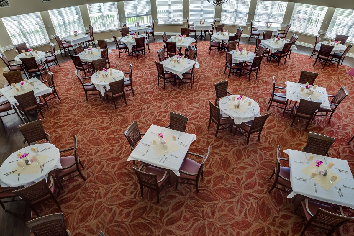 An empty dining area with round tables and chairs, set on a red patterned carpet with large windows in the background.
