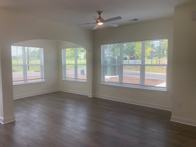 An empty room with large windows and a ceiling fan, featuring hardwood floors and ample natural light.