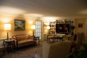 A cozy living room with a sofa, lamps, and a large television, decorated with artwork and books on shelves.