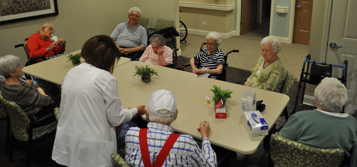 A group of elderly individuals gathered around a table with a caregiver in a nursing home setting.