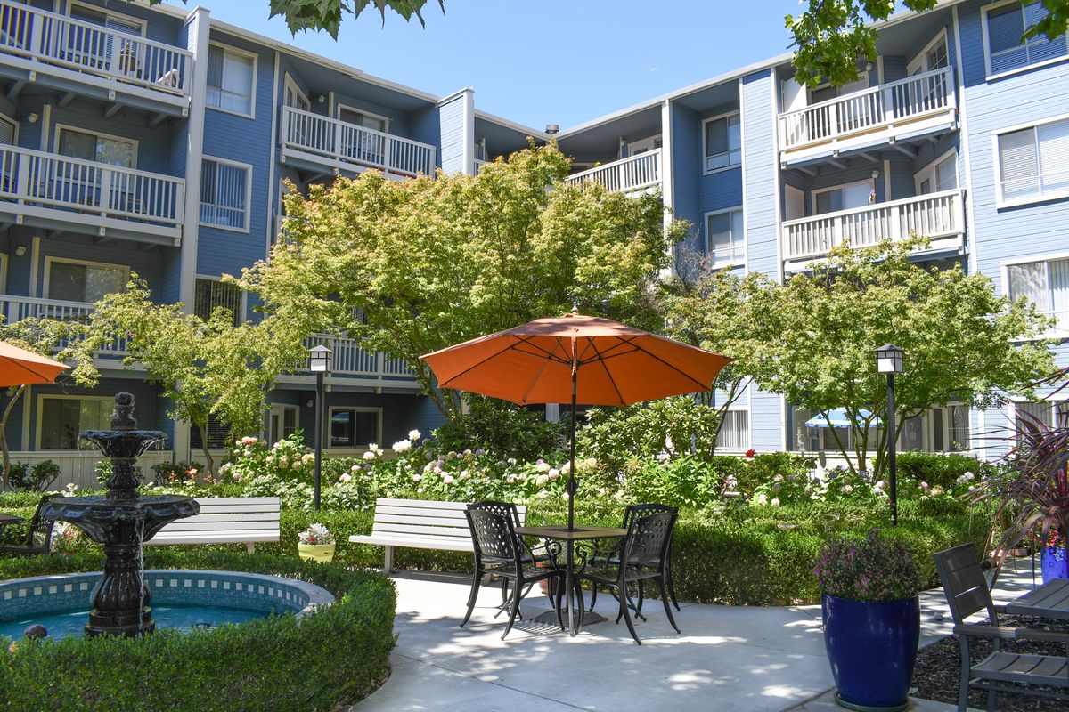 A serene apartment courtyard featuring a patio table with an orange umbrella, surrounded by lush greenery and benches.