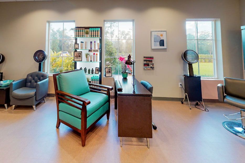 A cozy and inviting salon interior with a desk, teal armchair, and large windows.