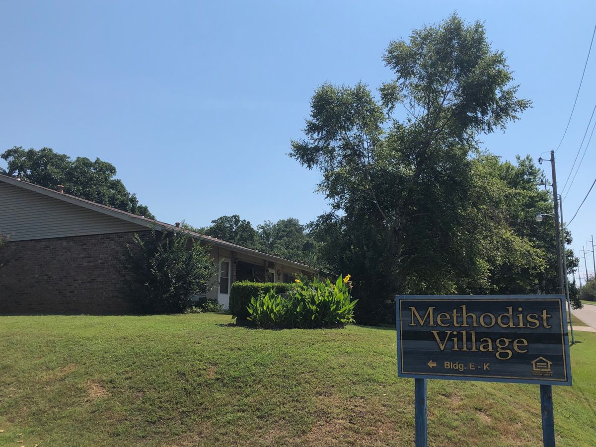 A building with a sign reading 'Methodist Village' surrounded by grass and trees under a clear sky.