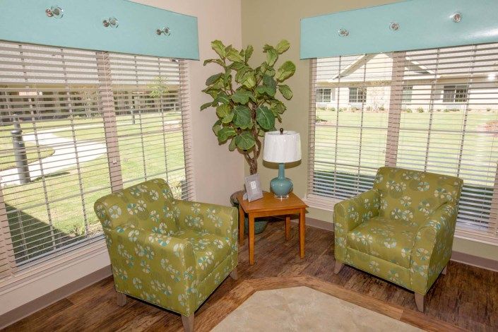 A cozy living room corner with two green patterned chairs, a small wooden table with a lamp, and a potted plant by the windows.