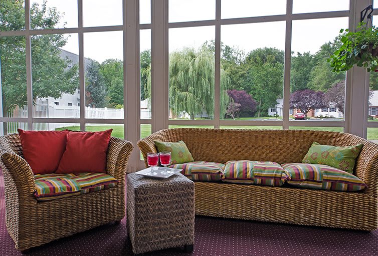 A cozy sunroom featuring wicker furniture and colorful cushions, with large windows offering a scenic outdoor view.