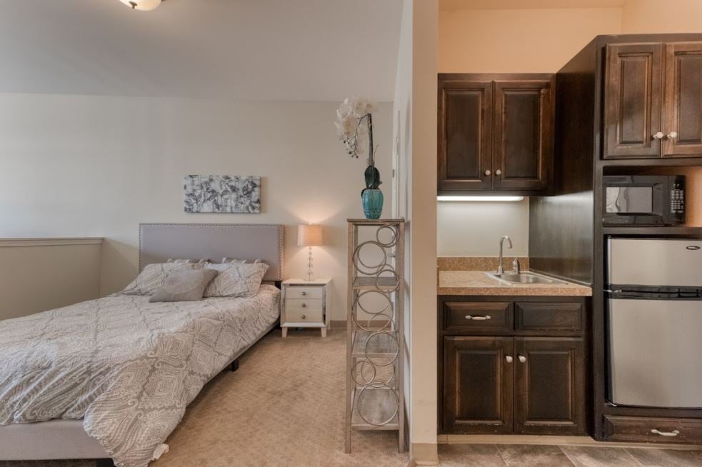 A cozy bedroom with adjacent kitchenette, featuring a comfortable bed, decorative lamp, and stylish cabinetry.