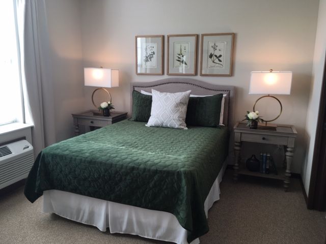 A cozy bedroom featuring a bed with green bedding, flanked by matching side tables and lamps, adorned with elegant wall art.