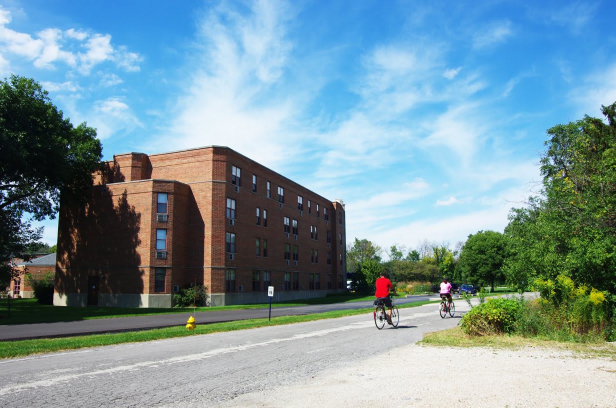 Two cyclists ride past a brick building under a bright blue sky.