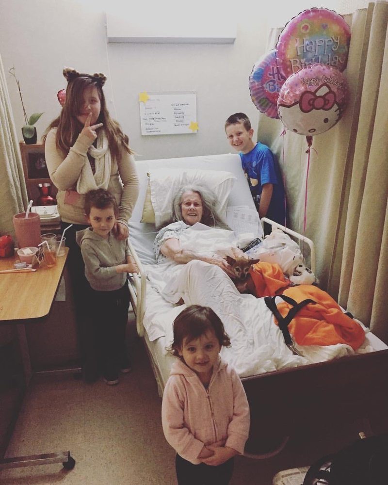 A family gathers around a hospital bed, with balloons in the background and children smiling.