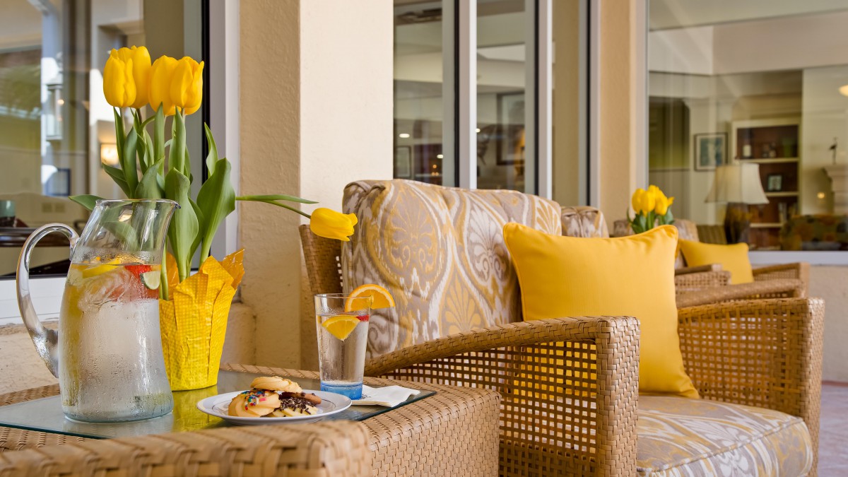 A cozy patio setup with wicker furniture, bright yellow cushions, a plate of food, and a refreshing drink beside a vase of yellow tulips.