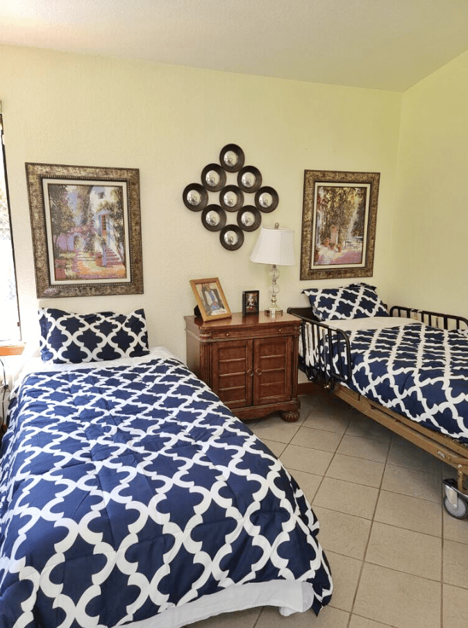 A cozy bedroom featuring two twin beds with blue and white patterned bedding, complemented by decorative elements such as framed artwork and a wooden nightstand with a lamp.