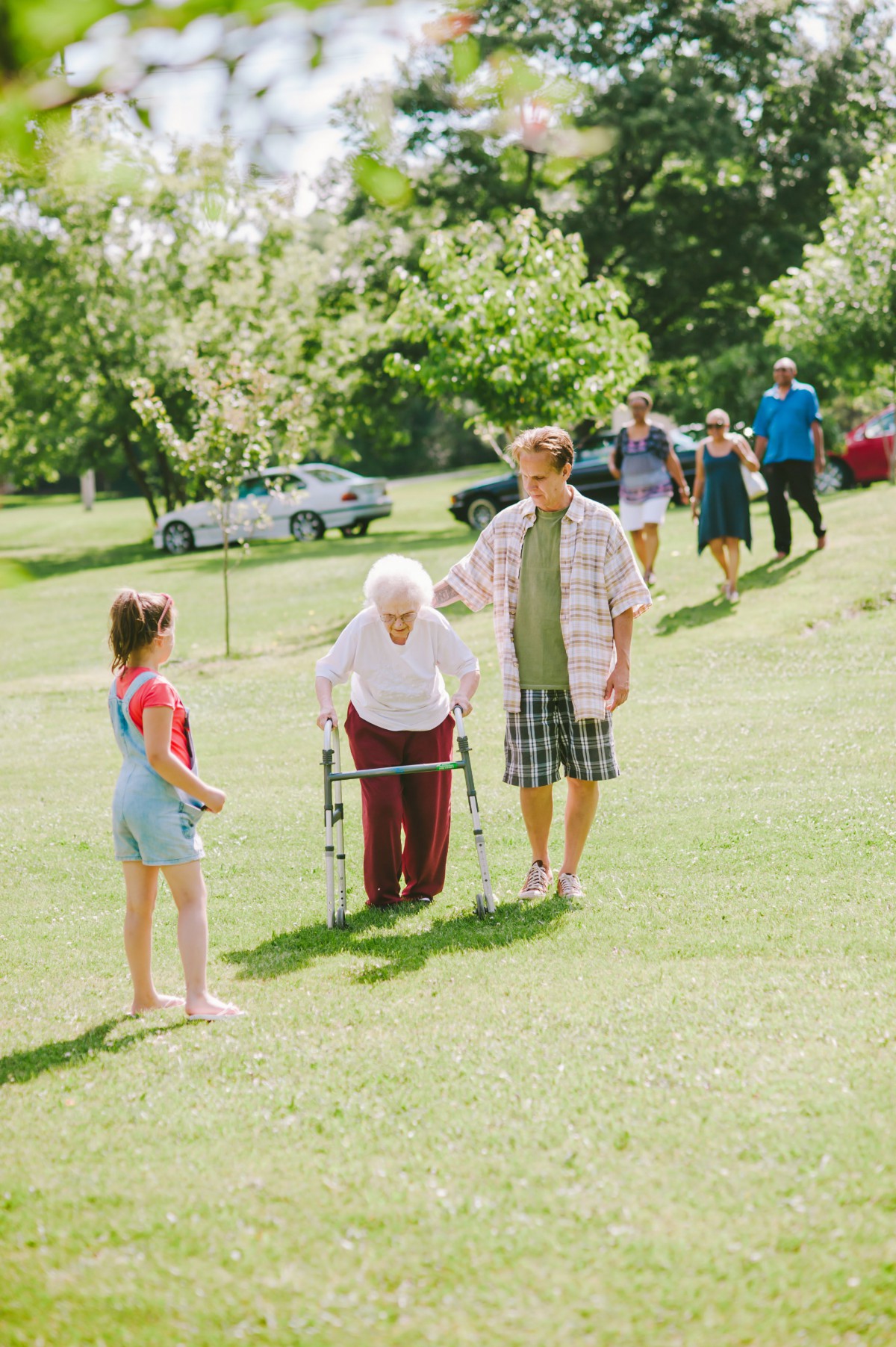 A family enjoying a sunny day in the park, with an elderly person using a walker and receiving assistance.