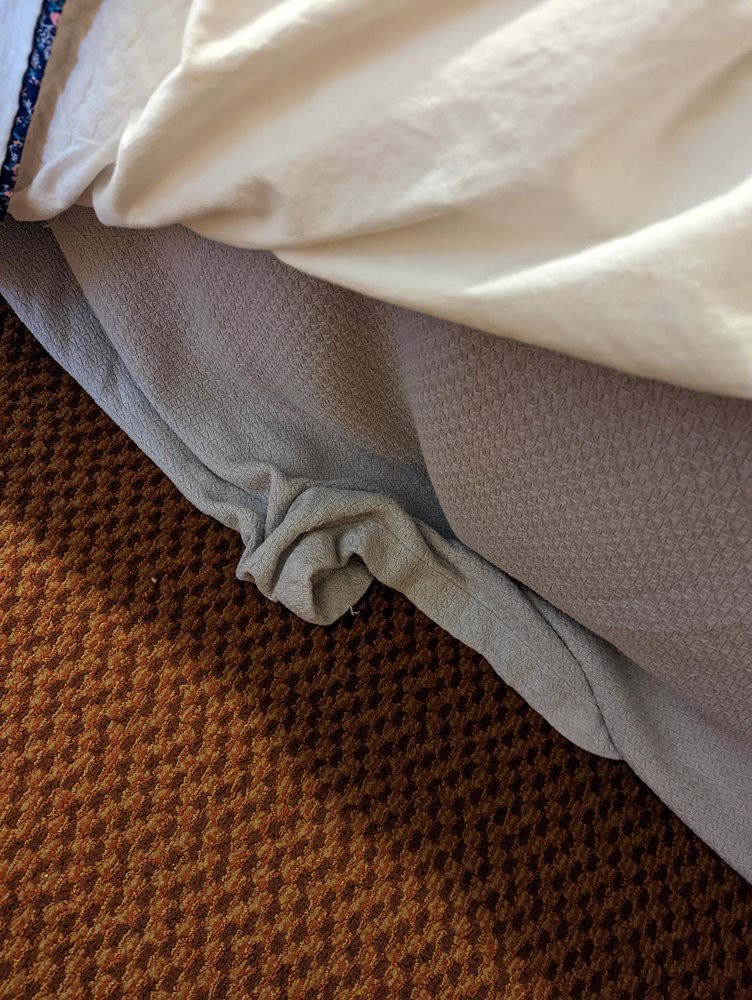 A close-up view of a bed with wrinkled sheets partially covering a mattress on a carpeted floor.