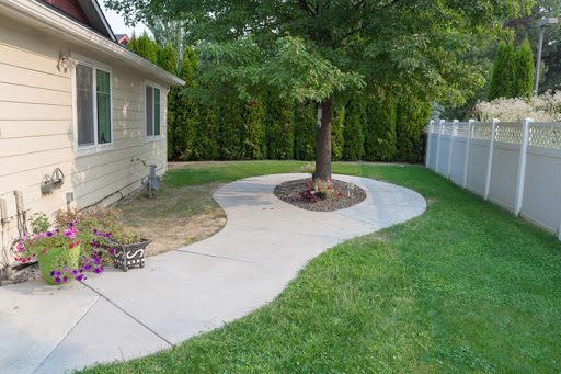A winding concrete pathway leading through a well-groomed garden with a tree and lush green grass.
