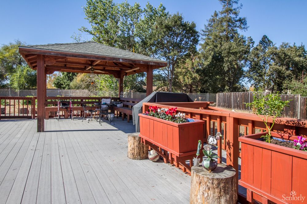 A spacious outdoor deck featuring a gazebo, grill, and seating area surrounded by planters and trees.