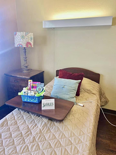 A cozy hospital room featuring a neatly made single bed with decorative pillows, a nightstand adorned with a lamp, and a tray of items.