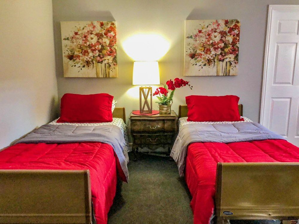 A cozy bedroom with twin beds adorned in red bedding, accompanied by a nightstand and decorative floral paintings above.