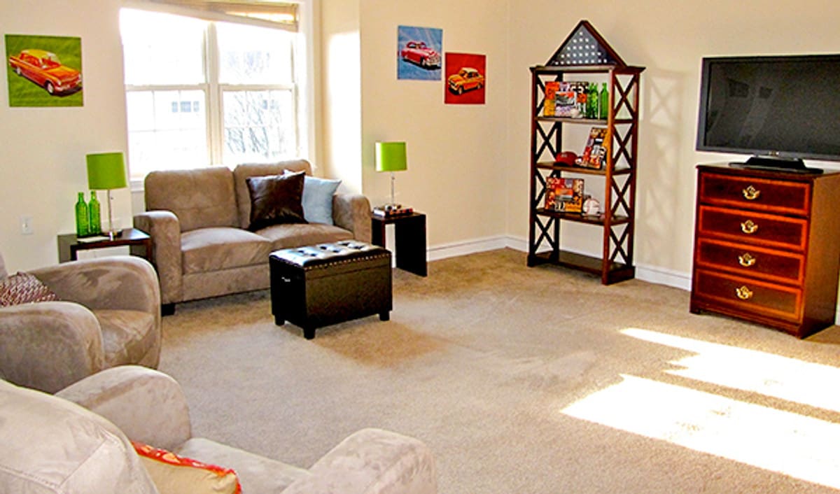 A cozy living room with a sofa, coffee table, bookshelf, and bright natural light from large windows.