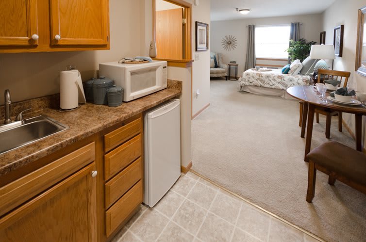 A cozy apartment kitchenette with wooden cabinets, featuring a microwave and refrigerator, leading into a living space.