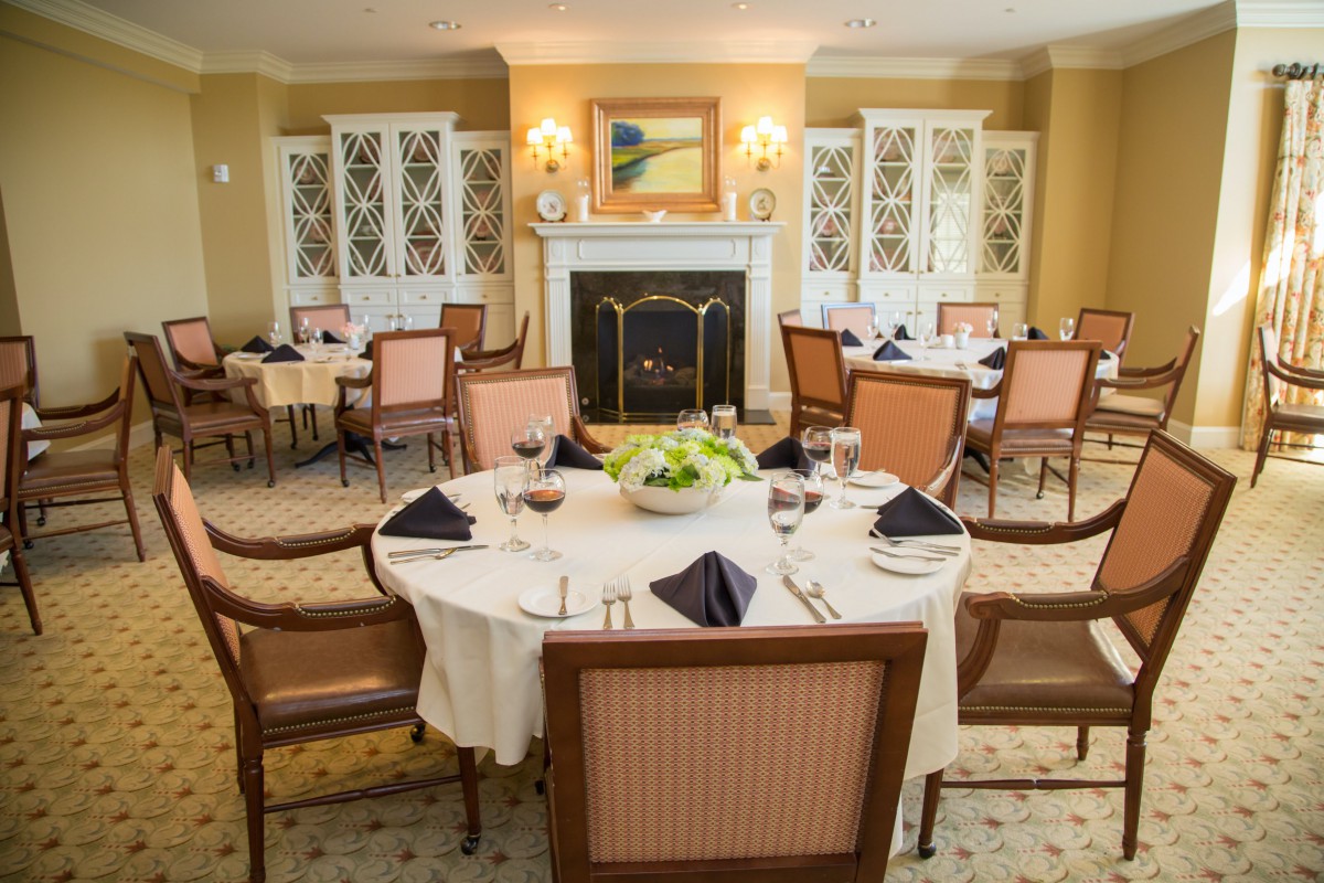 An elegantly set dining room with round tables, upholstered chairs, and a fireplace.