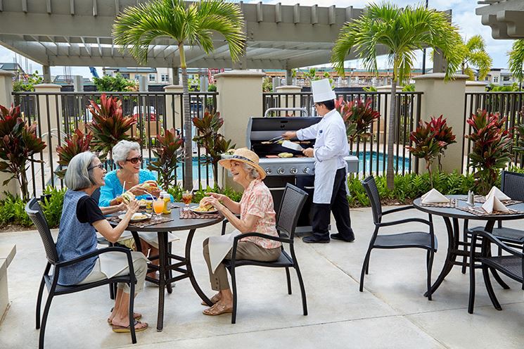 A group of seniors enjoys an outdoor meal on a patio, while a chef grills nearby.