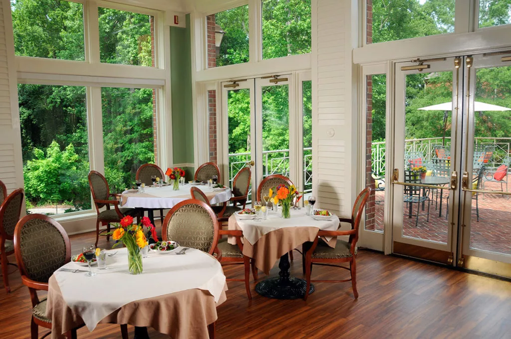 Elegant restaurant interior with tables and chairs, adorned with flowers and natural light streaming through large windows.