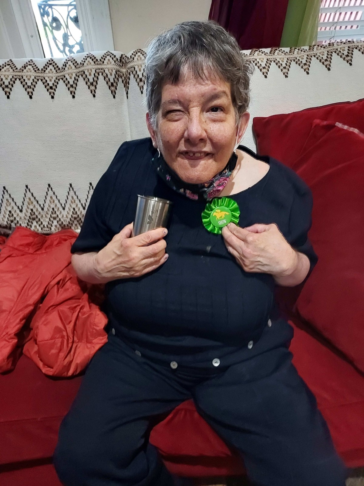 An elderly woman smiling while seated on a red couch, wearing a birthday badge.