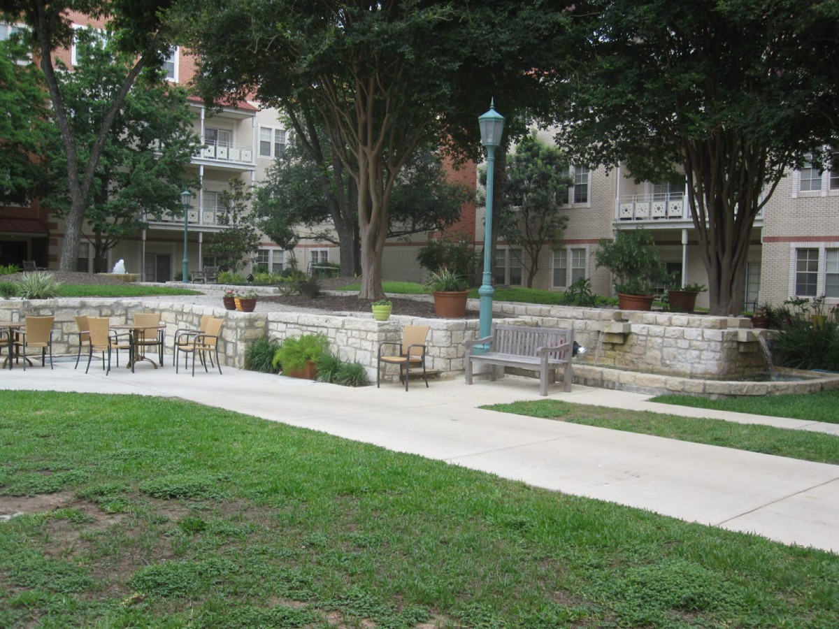 A serene park area featuring a pathway, benches, and chairs surrounded by greenery and trees.