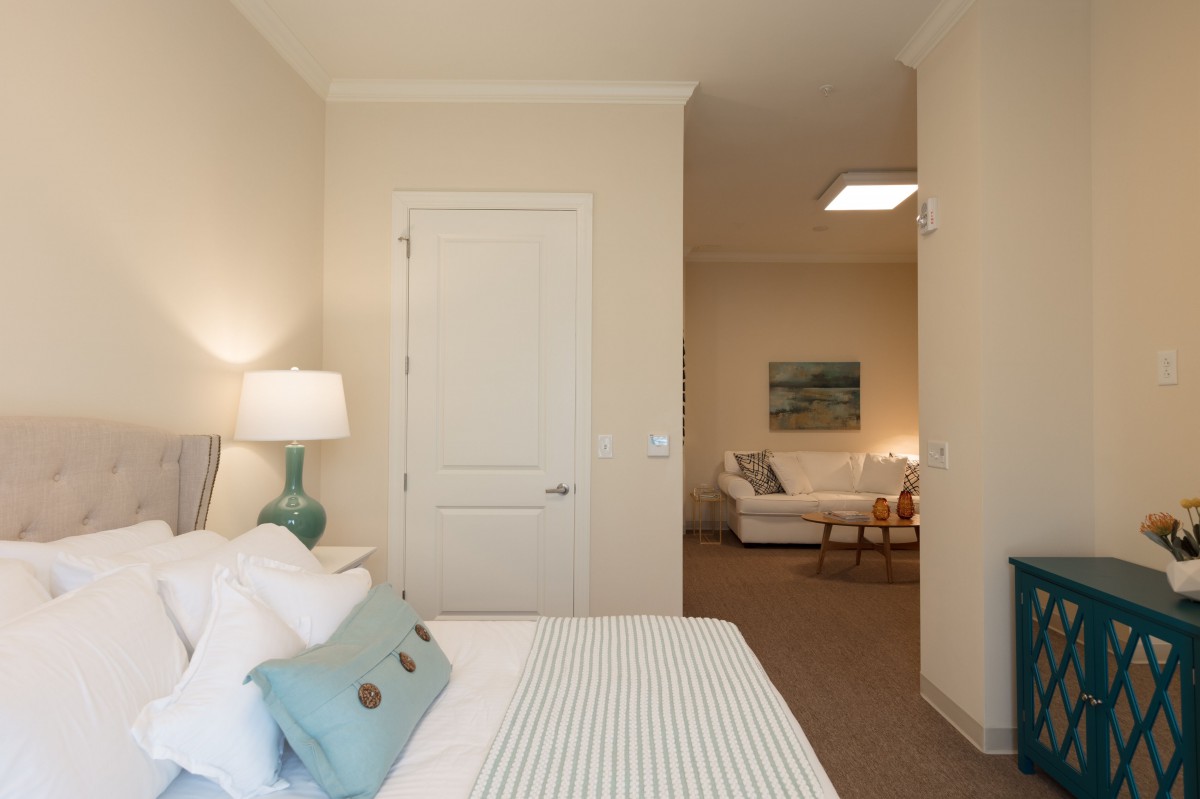 A cozy modern bedroom with a minimalist design, featuring a bed, bedside lamp, and view into a living area.