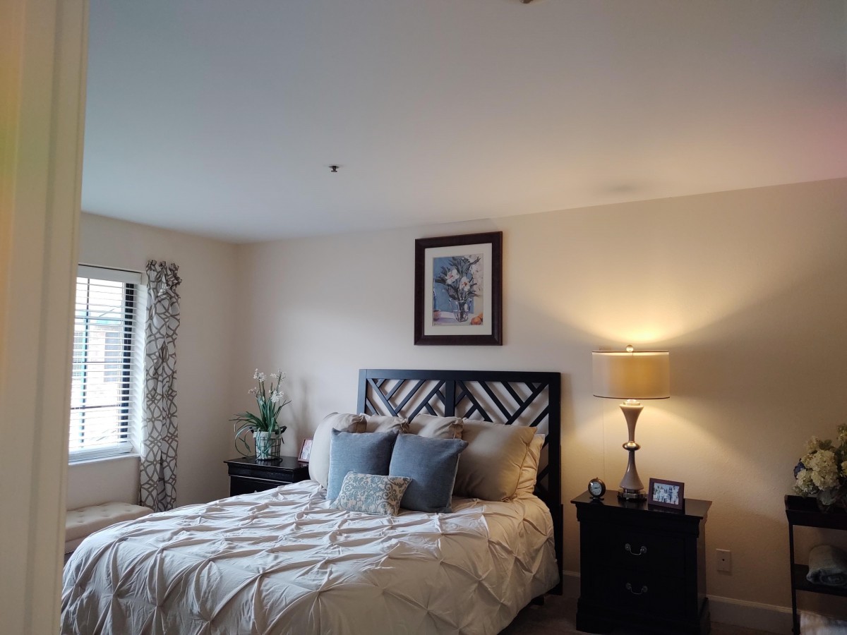 A cozy bedroom with a neatly made bed, accented by decorative pillows, a nightstand with a lamp, and artwork above the headboard.