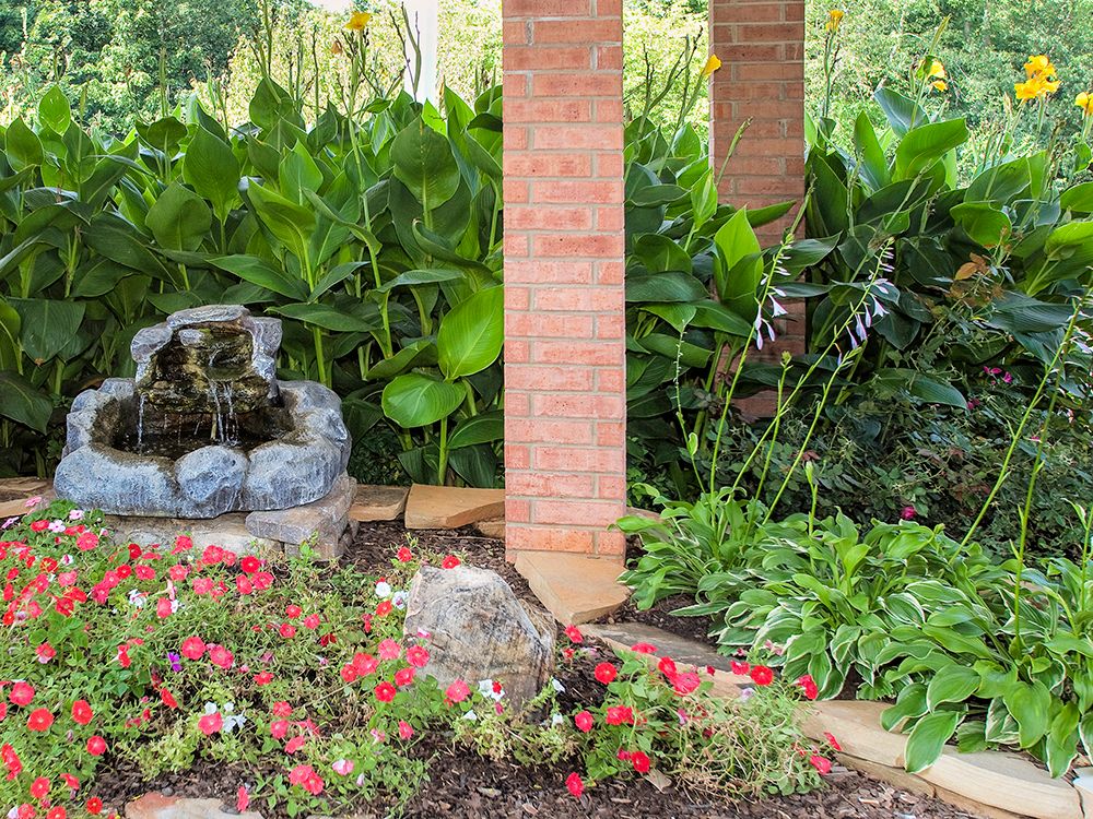 A tranquil garden scene featuring vibrant flowers, a small water fountain, and lush greenery surrounding a brick pillar.