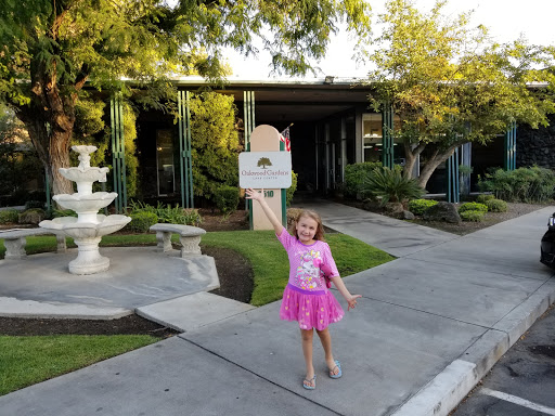 A cheerful child in a pink dress holding up a sign near a fountain on a sunny day.