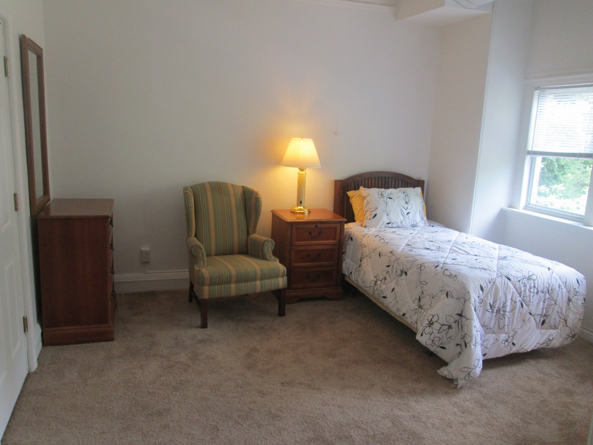 A simple bedroom with a single bed, nightstand, lamp, chair, and dresser.