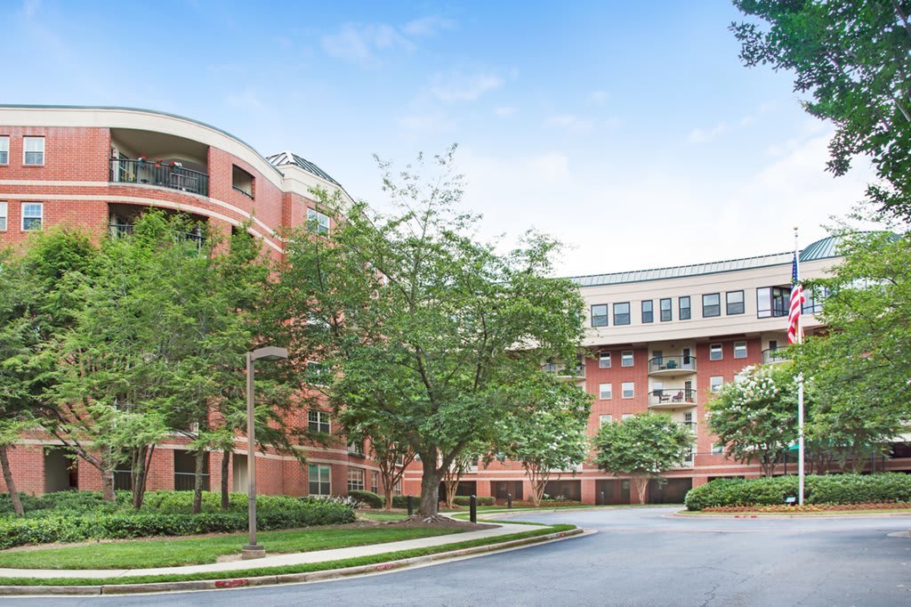 A view of a brick building surrounded by trees with a parking lot in the foreground on a clear day.