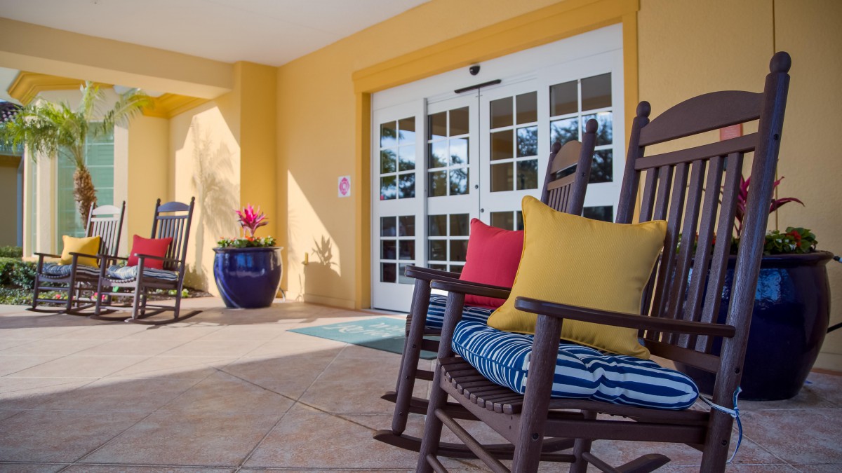 Colorful rocking chairs with cushions adorn a sunny porch entrance, complete with a potted plant.