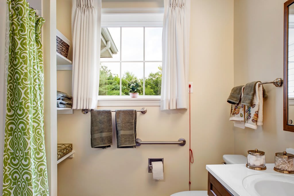 A small bathroom with a window, towels, toilet paper, and a handrail by the toilet.