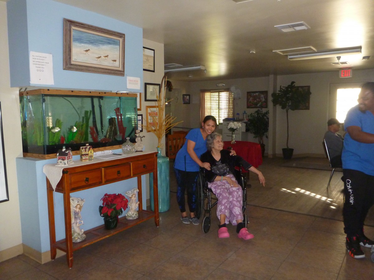A nurse standing beside an elderly woman in a wheelchair in a care facility's living area.