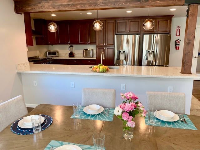 A modern kitchen and dining area featuring wooden cabinets, stainless steel appliances, and a spacious island with pendant lighting. The dining table is set with flowers as a centerpiece.