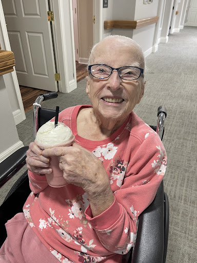 An elderly woman in a pink sweater enjoys a milkshake while smiling in her wheelchair.