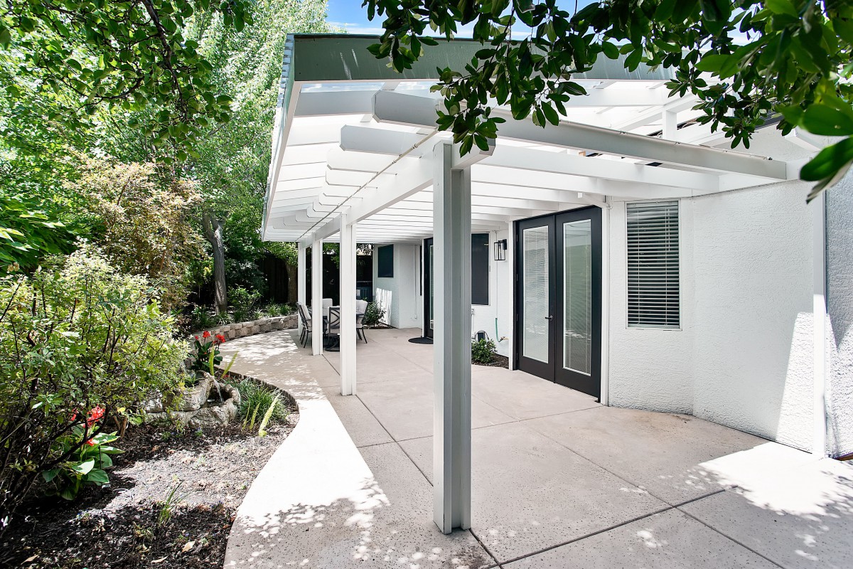 A modern patio with a white pergola, surrounded by lush greenery.